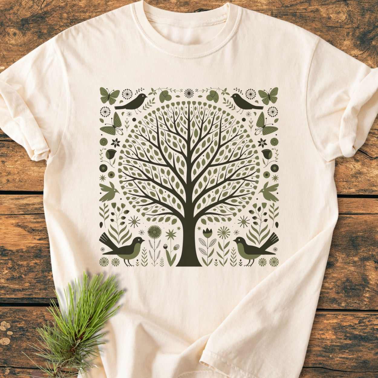 Nature Love - Vintage Wash Tee featuring a stylized tree with folk-art motifs on a cream-colored T-shirt, laid on a wooden surface, with a pine twig beside it.