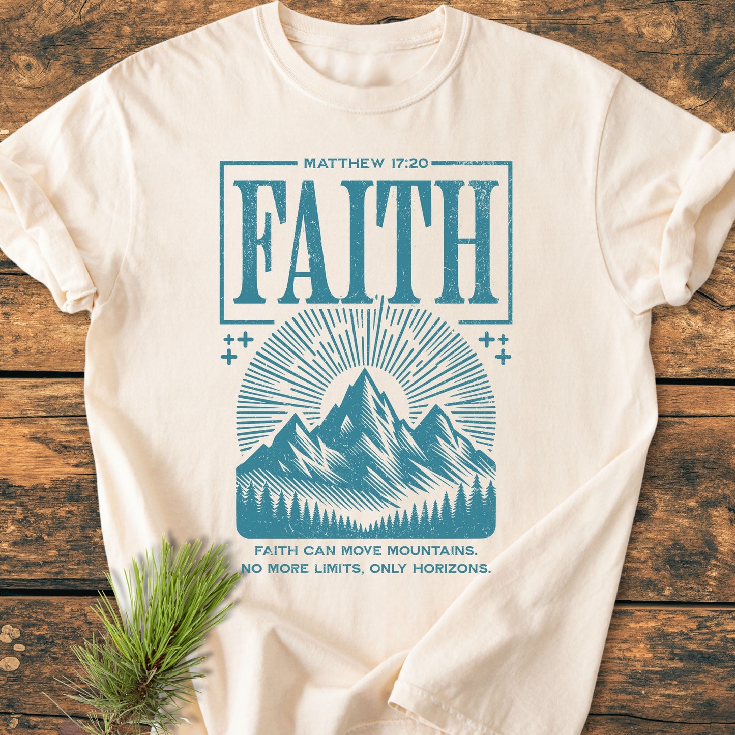 Faith Moves Mountains - Vintage Wash Tee: Cream-colored T-shirt with teal mountain graphic and Faith slogan, laid on rustic wood. Includes biblical reference and motivational text. Pine sprig accents the presentation.