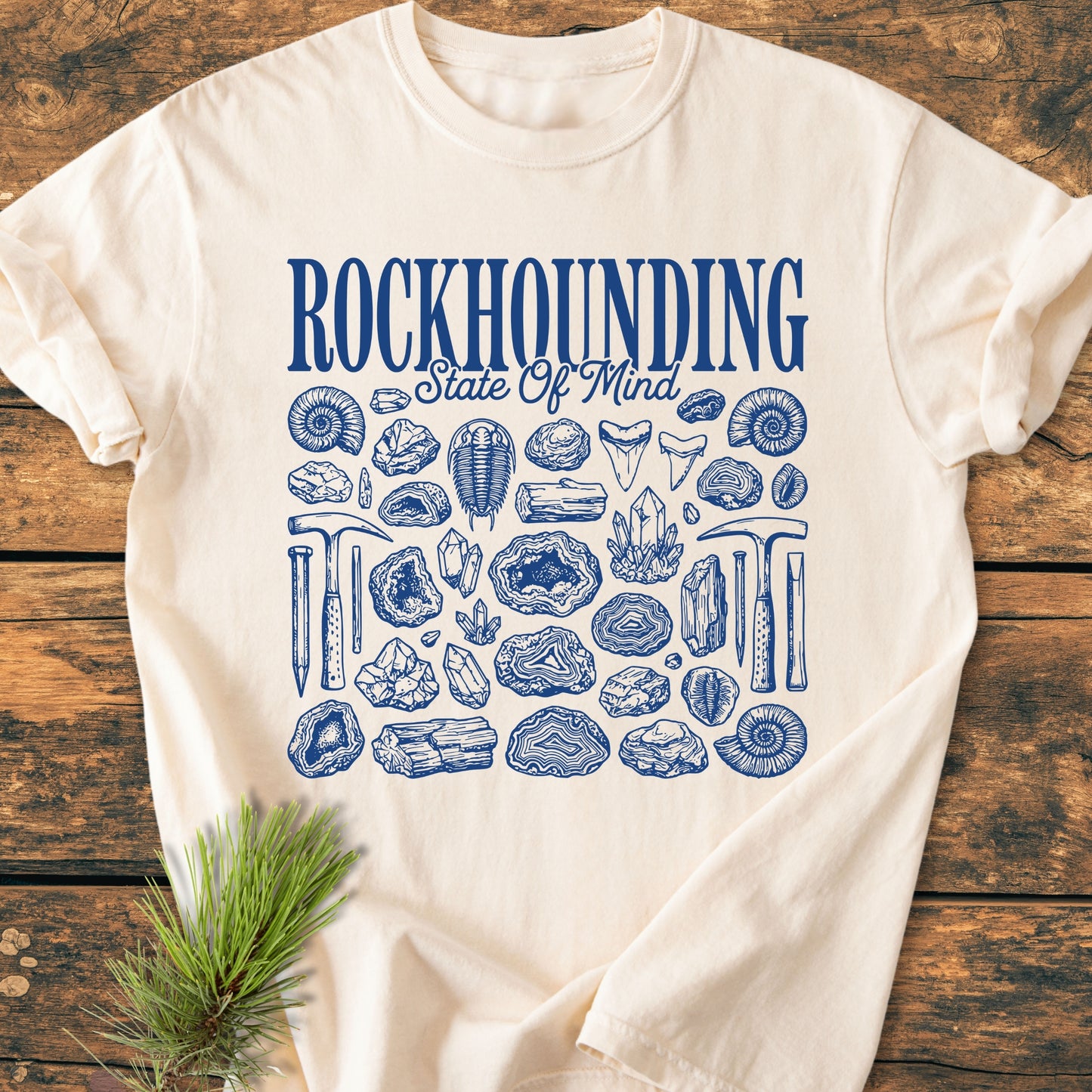 Rockhounding State Of Mind - Vintage Wash Tee features a bold blue graphic and detailed illustrations of fossils, minerals, and tools, emphasizing a rock-collecting theme on a light-colored, laid-flat T-shirt.
