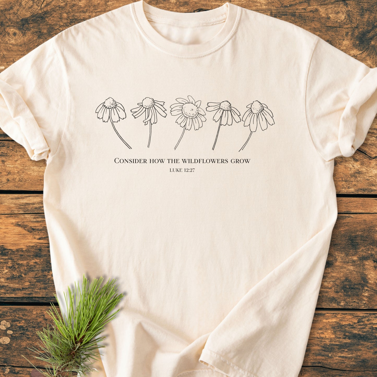Consider How The Wildflowers Grow - Vintage Wash Tee featuring a black line-drawing of wildflowers and text from Luke 12:27 on a cream-colored T-shirt, laid flat on rustic wood.