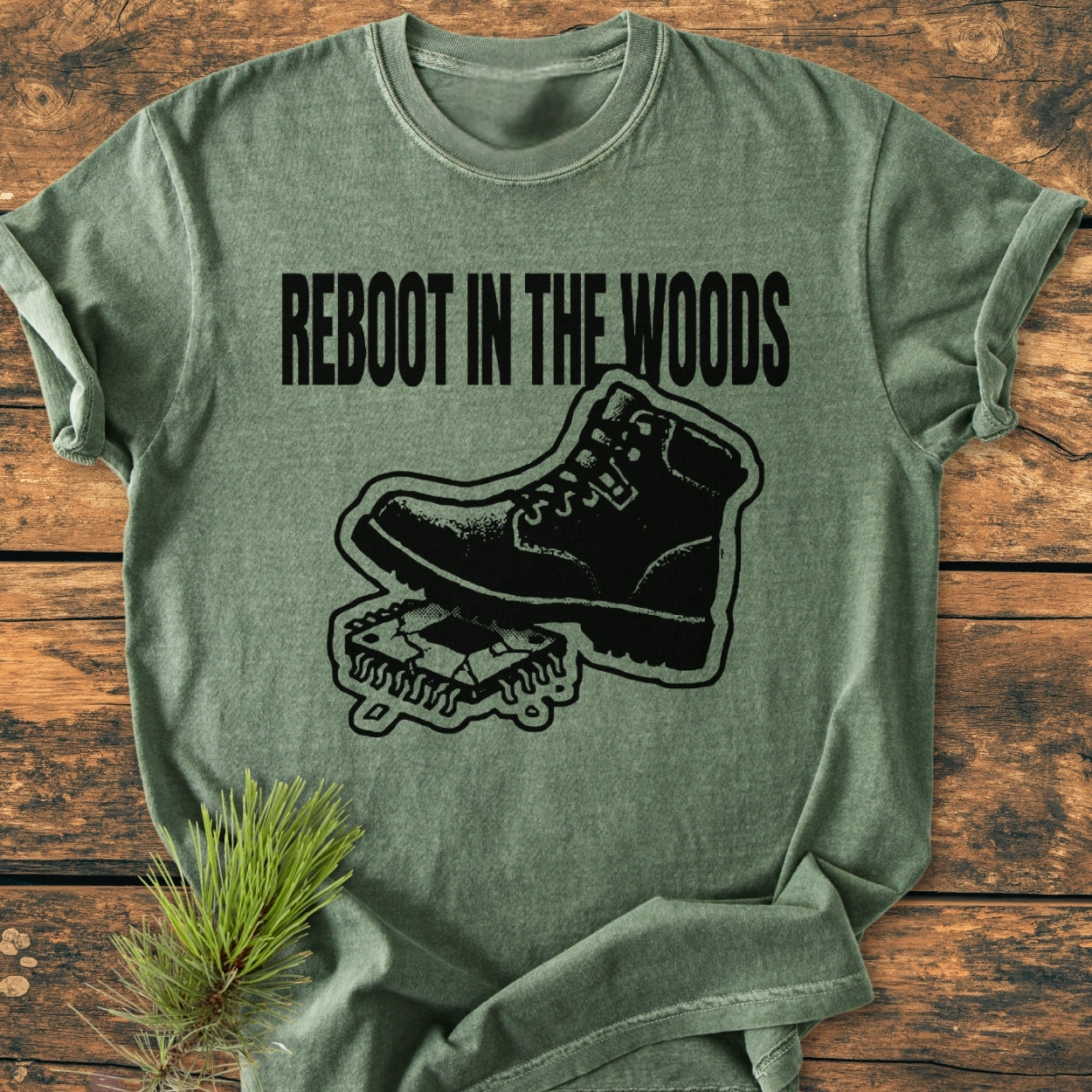 Reboot In The Wood - Vintage Wash Tee features a heather-green T-shirt with bold REBOOT IN THE WOODS text and a hiking boot on a circuit board graphic, highlighting its unique outdoorsy theme.