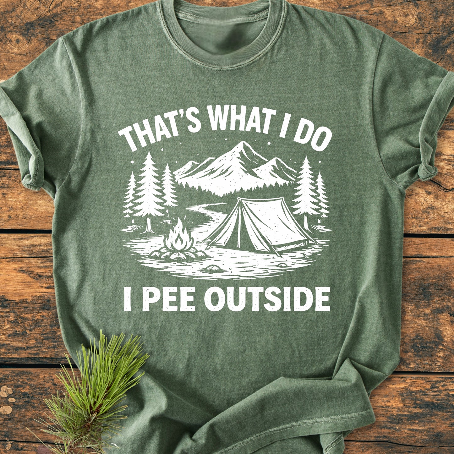 I Pee Outside - Vintage Wash Tee featuring a rustic camping scene with pine trees, mountains, a tent, and campfire, laid on a wooden surface. Made from soft, pre-shrunk, 100% ring-spun cotton.
