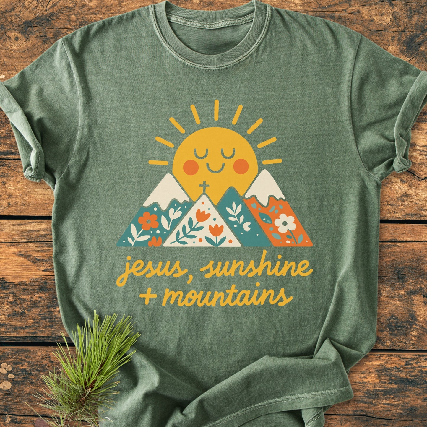 Jesus, Sunshine, Mountains - Vintage Heavyweight Tee: Green crew-neck T-shirt with a cheerful sun and mountain graphic, hand-lettered script, laid on wood with pine needles beside, highlighting comfort and durability.
