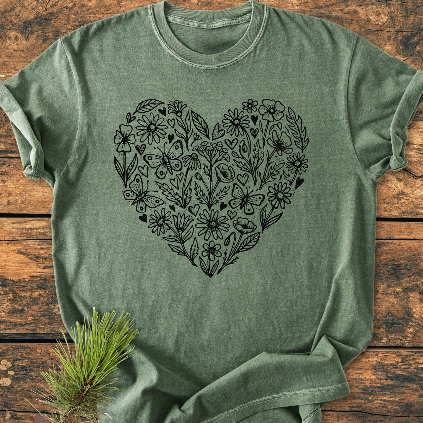 Wildflower Heart Butterfly Soul - Vintage Heavyweight Tee in heather-green, featuring a heart-shaped cluster of black line art flowers and butterflies, displayed flat on wood with a pine sprig accent.