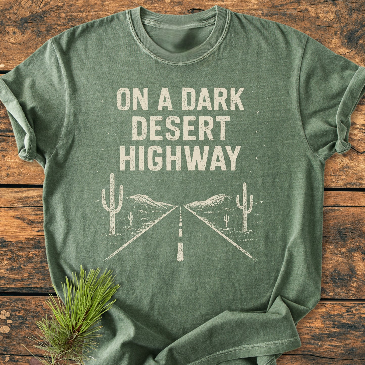 On A Dark Desert Highway - Vintage Wash Tee displayed on wood, featuring distressed text and desert line-drawing. Styled with pine sprig, highlighting its rugged, trail-ready appeal for hikers.