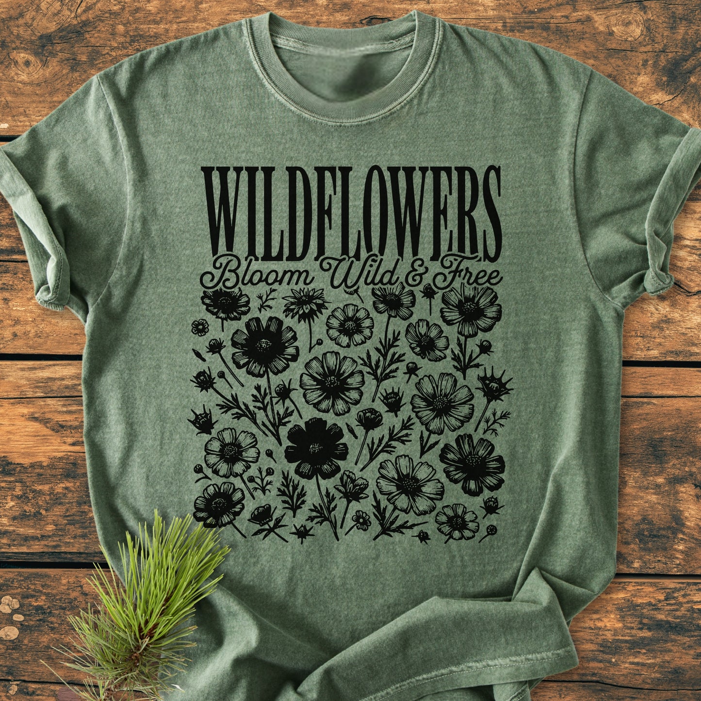 Wildflowers Bloom Wild And Free - Vintage Wash Tee featuring bold WILDFLOWERS text and black wildflower illustrations on a green short-sleeve shirt, laid on a wooden surface with a pine sprig beside it.