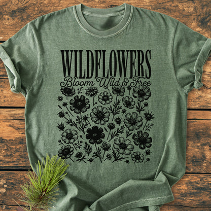 Wildflowers Bloom Wild And Free - Vintage Wash Tee featuring bold WILDFLOWERS text and black wildflower illustrations on a green short-sleeve shirt, laid on a wooden surface with a pine sprig beside it.