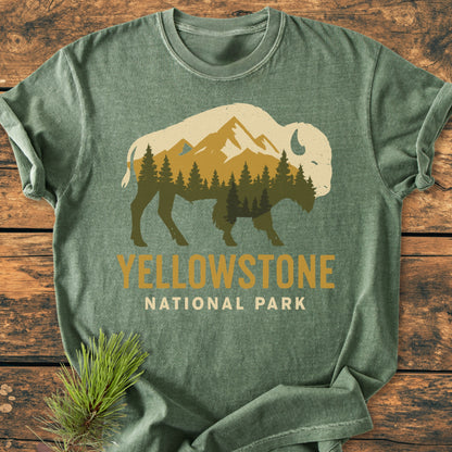 Yellowstone Bison - Vintage Wash Tee features a bison silhouette with a pine forest and mountain scene. YELLOWSTONE NATIONAL PARK text is below. Laid on wooden surface with pine sprig accent.
