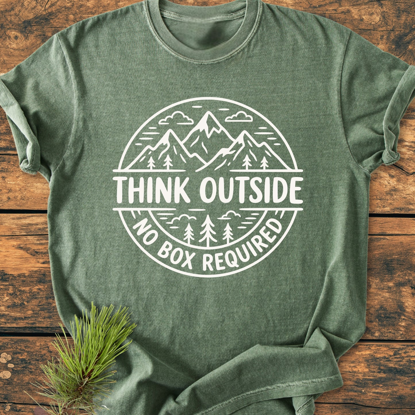 Heather-green Think Outside - Vintage Wash Tee with a white circular graphic of mountains and trees, laid on a wooden surface. The slogan reads, “THINK OUTSIDE, NO BOX REQUIRED.”