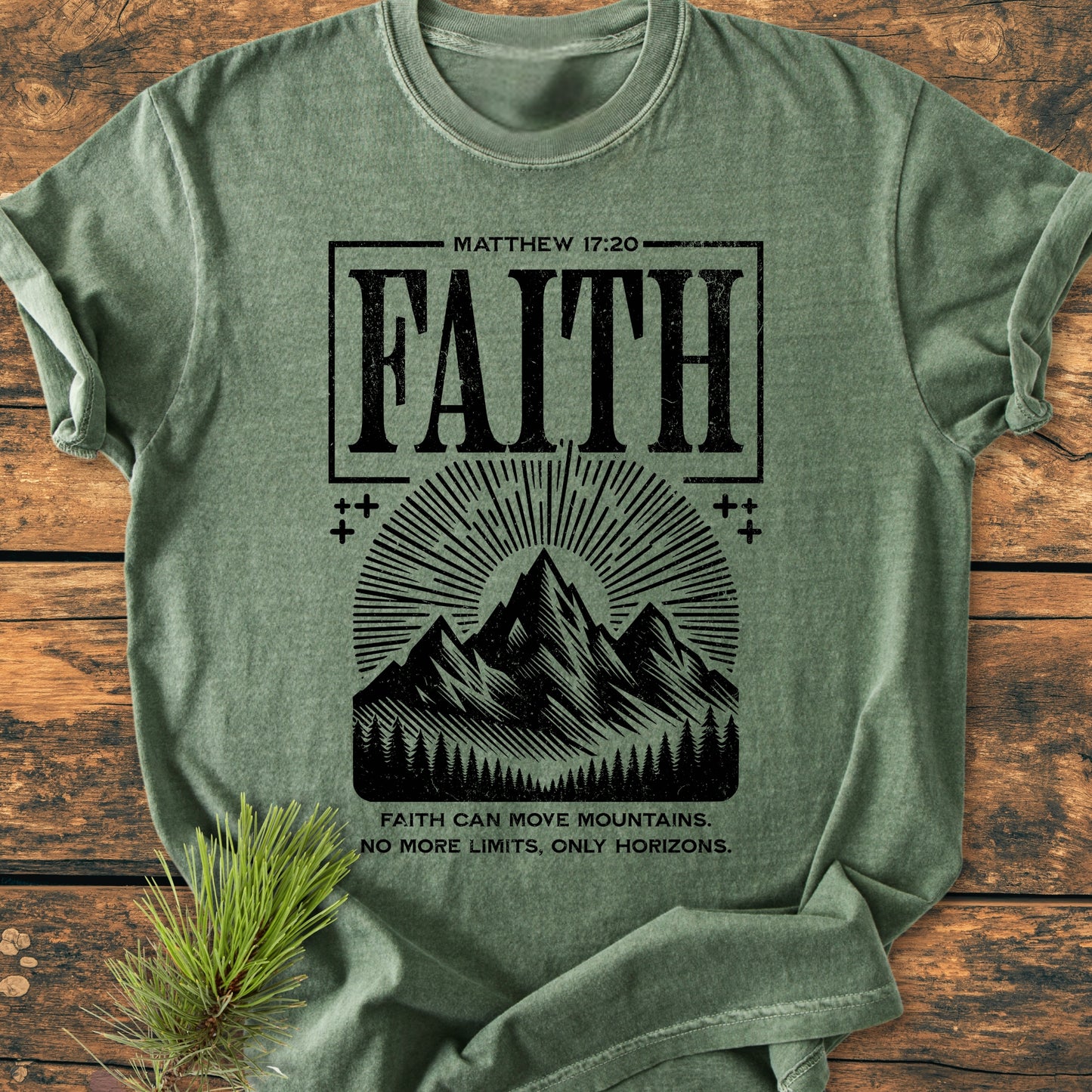 Faith Moves Mountains - Vintage Wash Tee features MATTHEW 17:20 and FAITH with a stylized mountain and pine tree graphic, laid flat on wood with a pine branch.