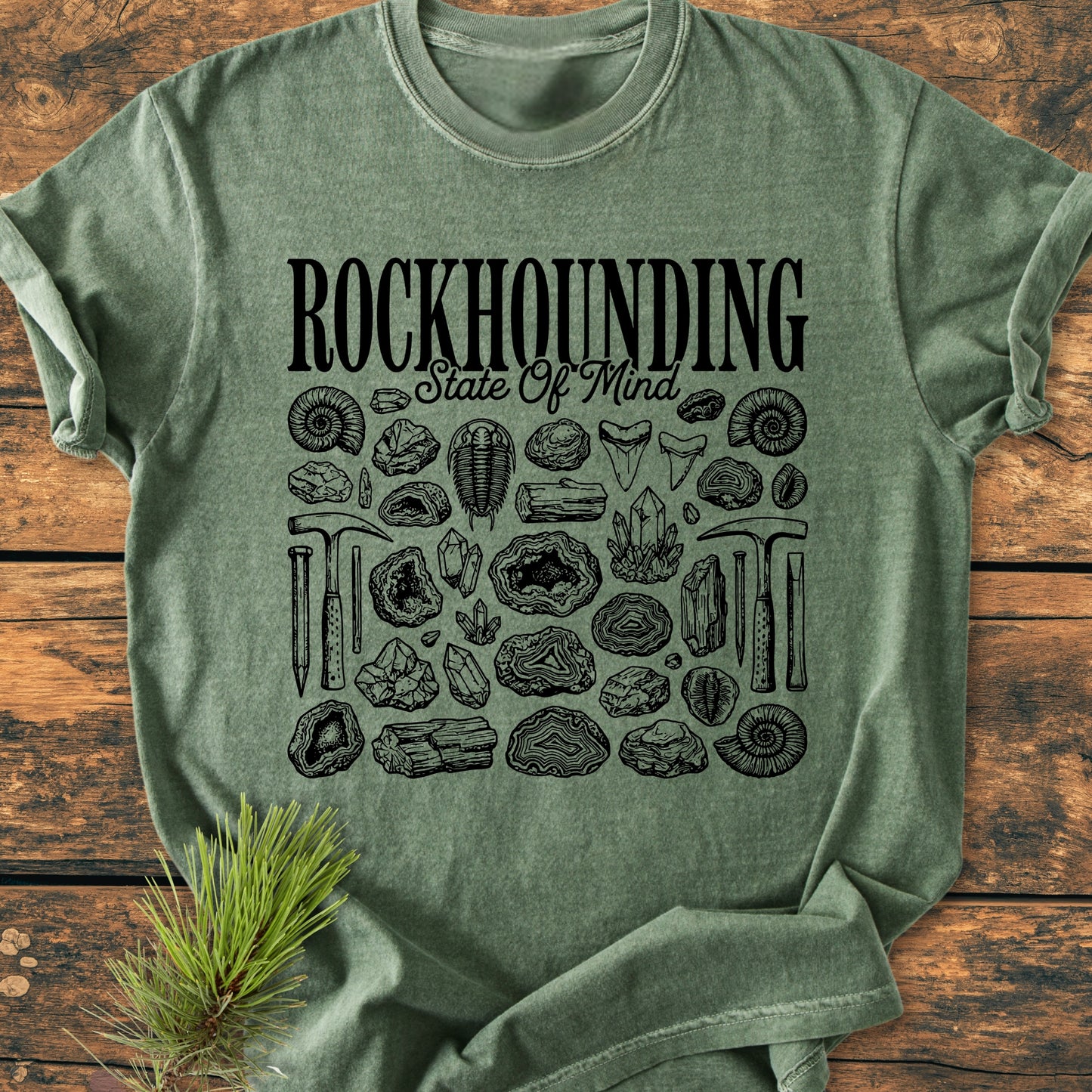 Rockhounding State Of Mind - Vintage Wash Tee featuring bold ROCKHOUNDING text, script State Of Mind, and detailed fossil and tool illustrations, laid on a wooden surface. Comfortable, durable, and pre-shrunk.