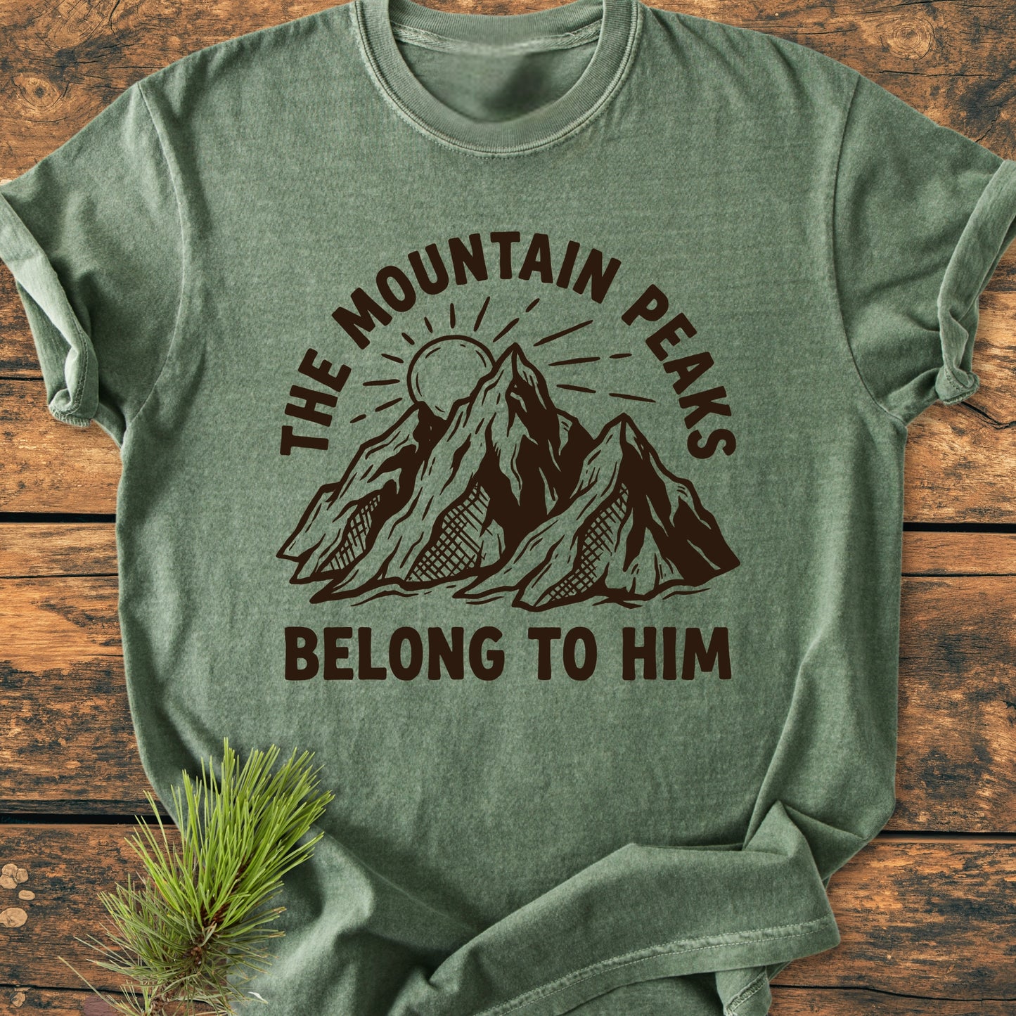 The Mountain Peaks Belong To Him - Vintage Wash Tee: Folded forest-green T-shirt featuring rugged mountain peaks graphic with sun, surrounded by text, on wooden surface with a pine sprig beside it.