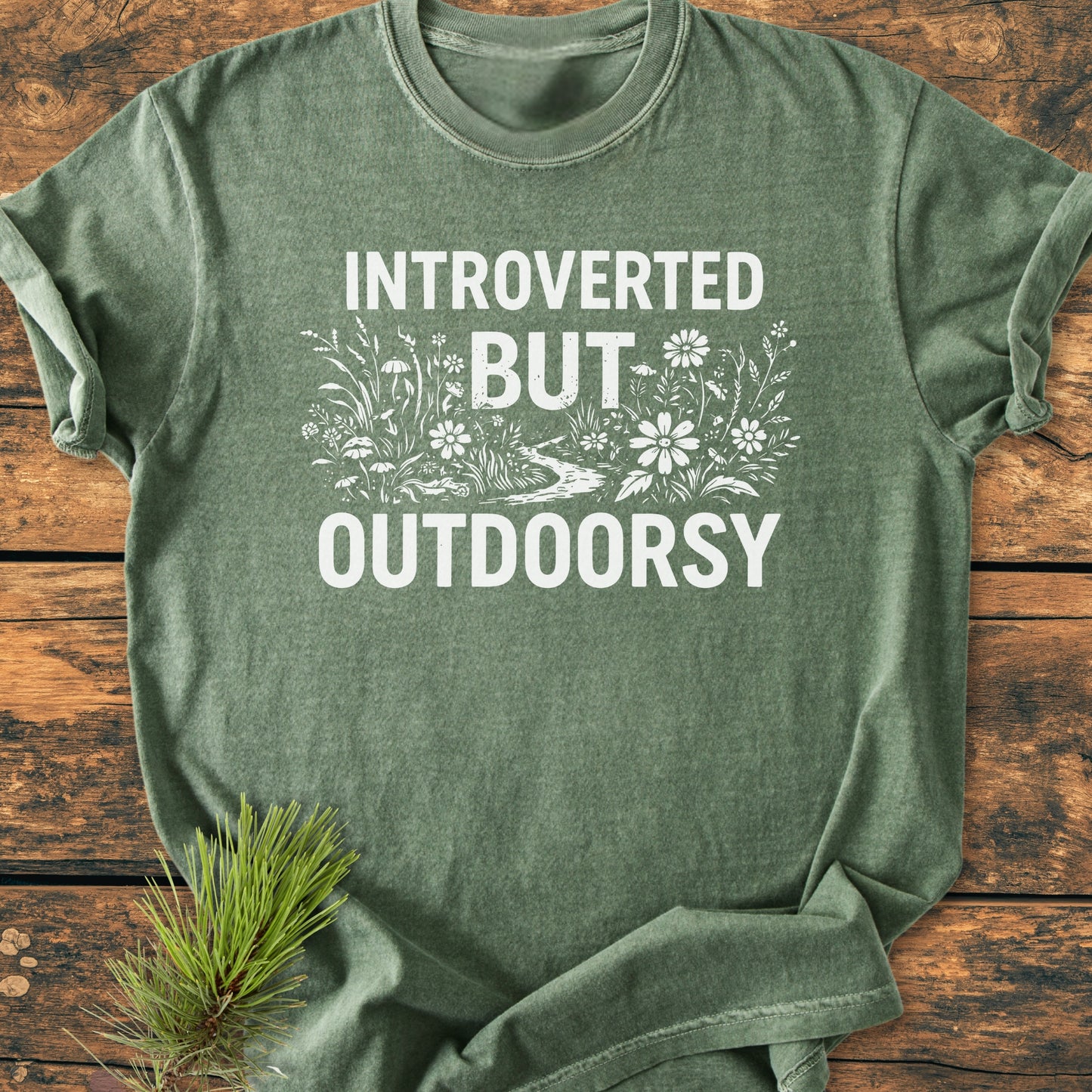 Introverted But Outdoorsy - Vintage Wash Tee: Green heather-style T-shirt with white graphic, featuring 'INTROVERTED BUT OUTDOORSY' and meadow motif. Displayed on wooden surface with pine needles for an outdoorsy touch.