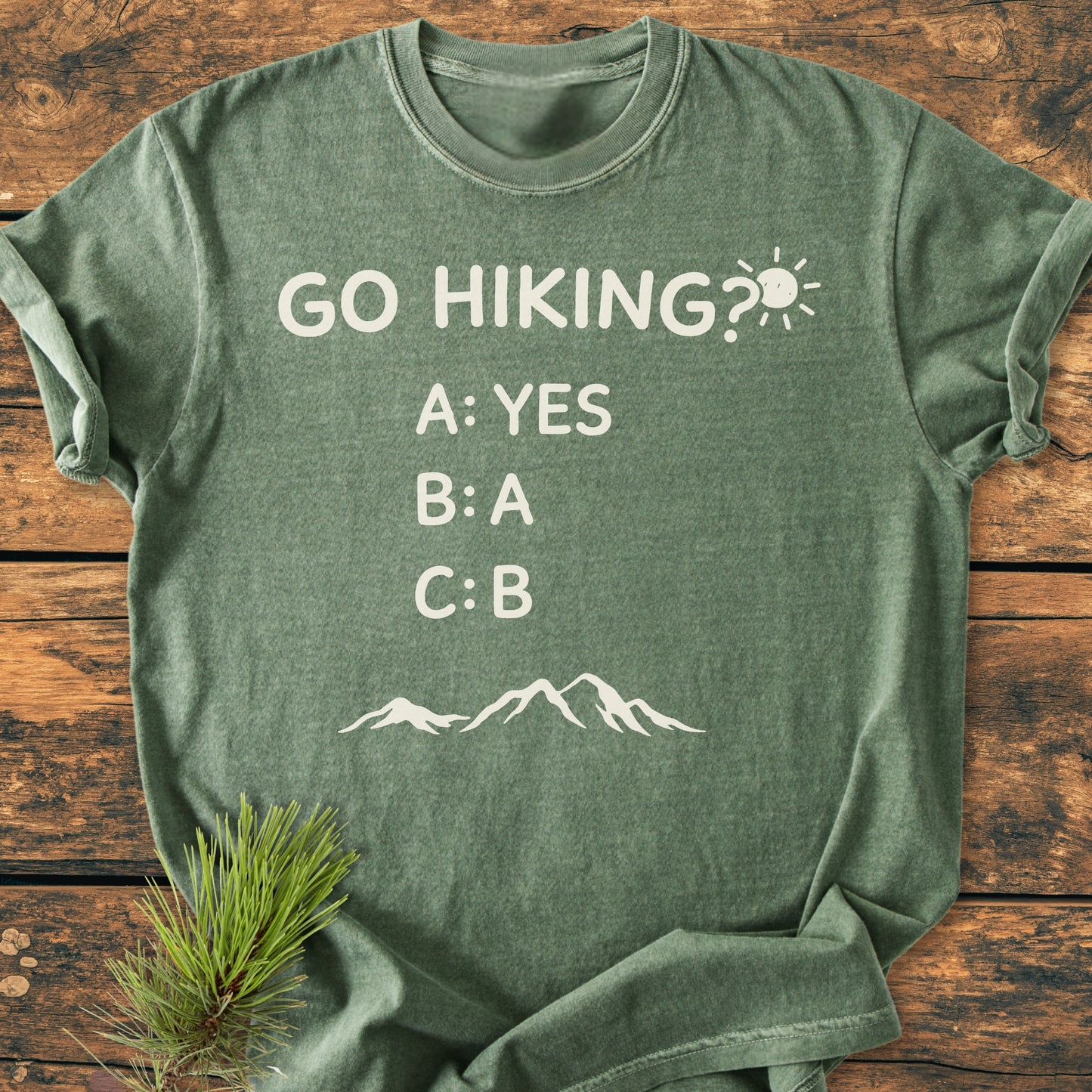 Go Hiking? - Vintage Wash Tee in heather-green, featuring playful text and mountain silhouette, styled with a pine branch, showcasing its relaxed fit and soft-washed, garment-dyed fabric.
