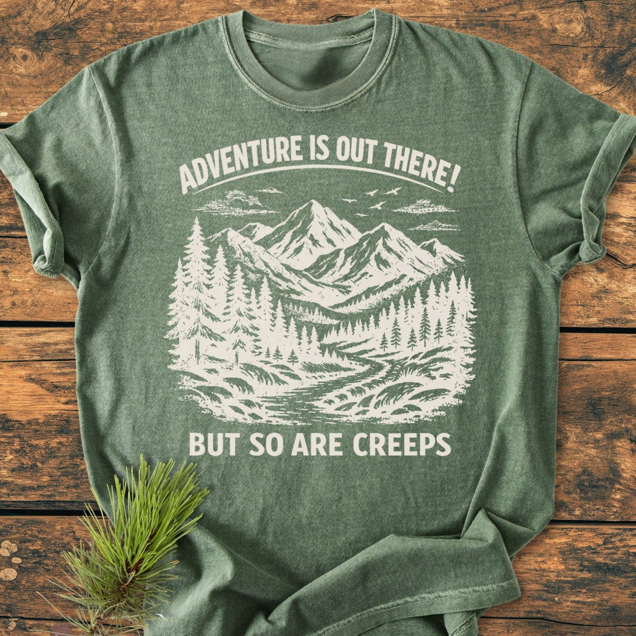 Adventure Is Out There - Vintage Wash Tee, featuring a mountain-and-forest scene with a witty slogan, laid flat on a wooden surface with a pine sprig accentuating its outdoorsy theme.