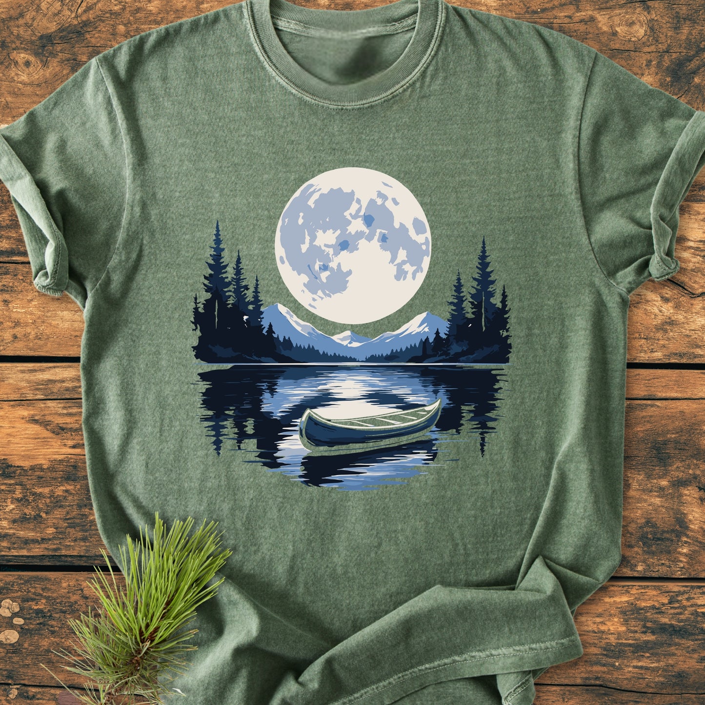 Moonlit Canoe - Vintage Wash Tee features a serene outdoor scene with a canoe on a lake, pine trees, mountains, and a full moon, laid out on a wooden surface with a pine sprig.
