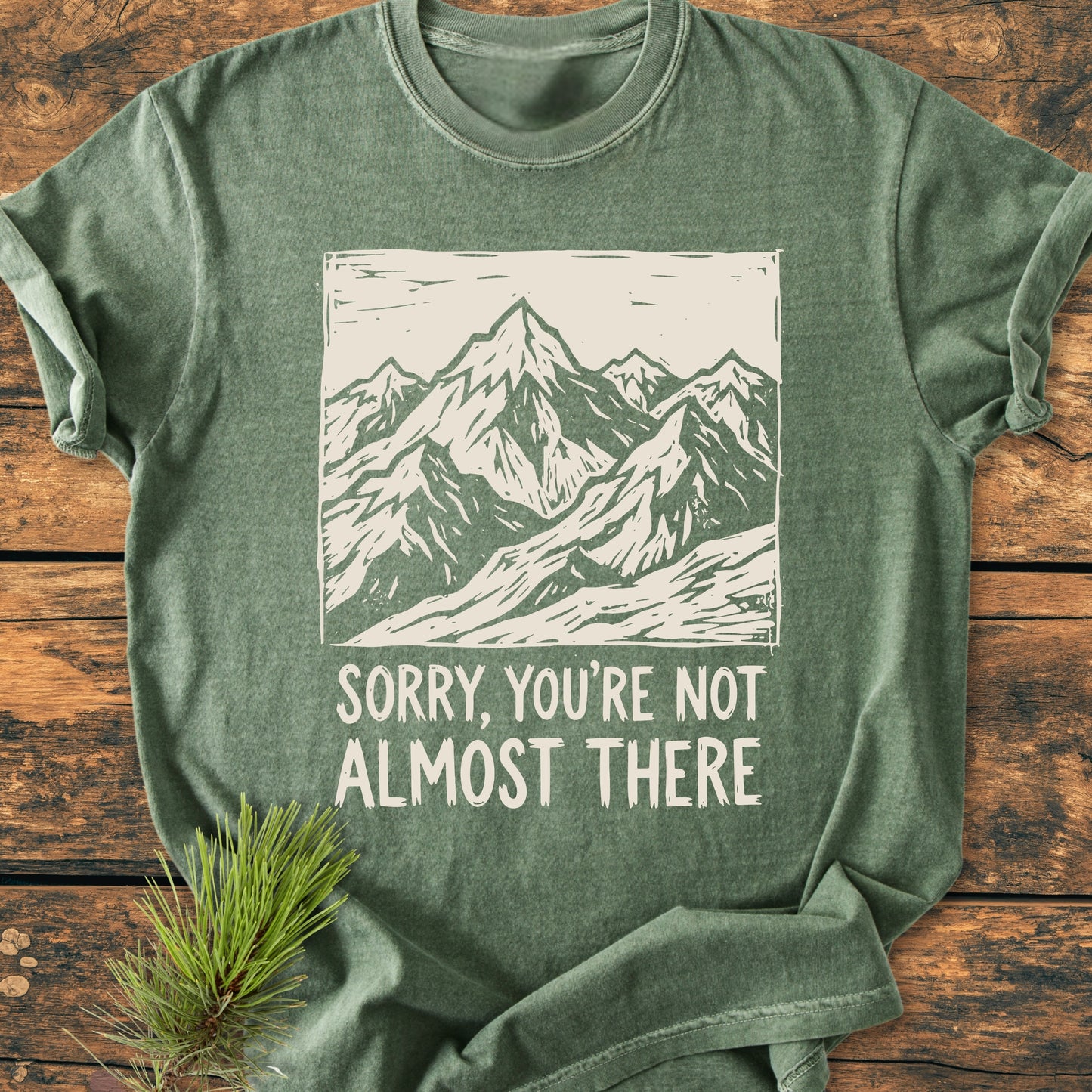 Not Almost There - Vintage Wash Tee displayed on wood, features a white mountain illustration with slogan. Pine sprig beside enhances the rustic appeal, highlighting the soft, garment-dyed cotton fabric.