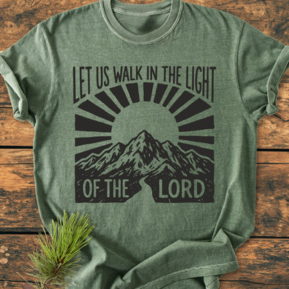 Let Us Walk In The Light Of The Lord - Vintage Wash Tee features a mountain graphic and sunburst design on a heather-green background, laid on wood with a pine sprig accentuating its rustic charm.