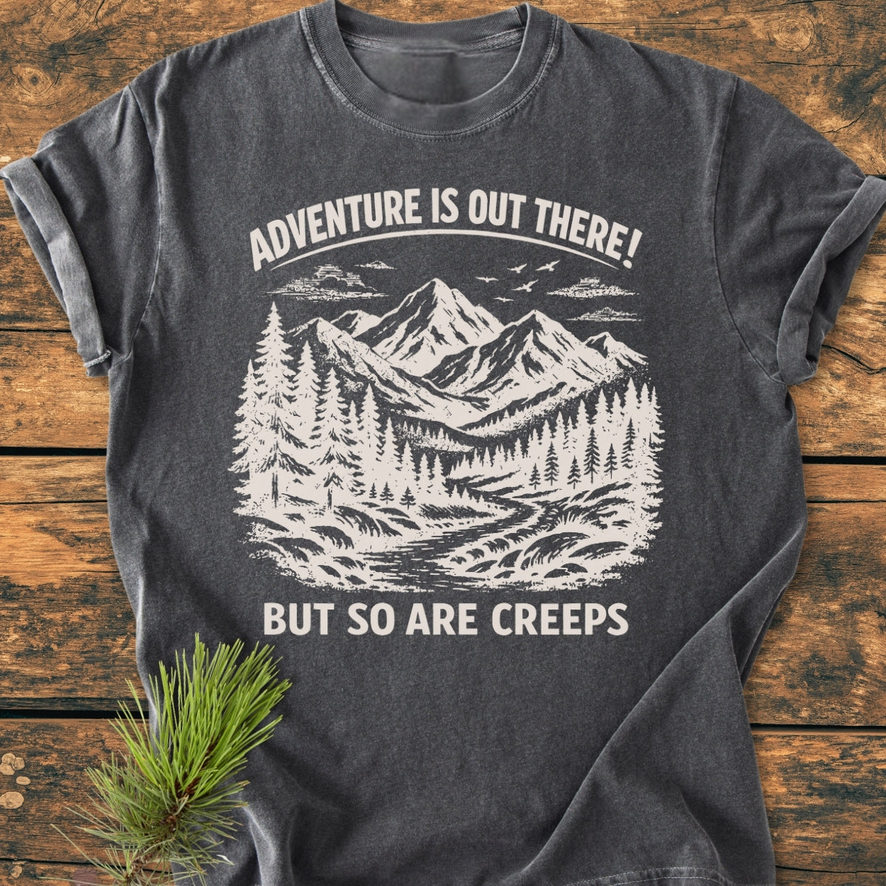 Adventure Is Out There - Vintage Wash Tee featuring a hand-drawn mountain landscape with trees and stream, laid on wood with pine sprig. Bold text adds a playful touch to this cozy, durable shirt.