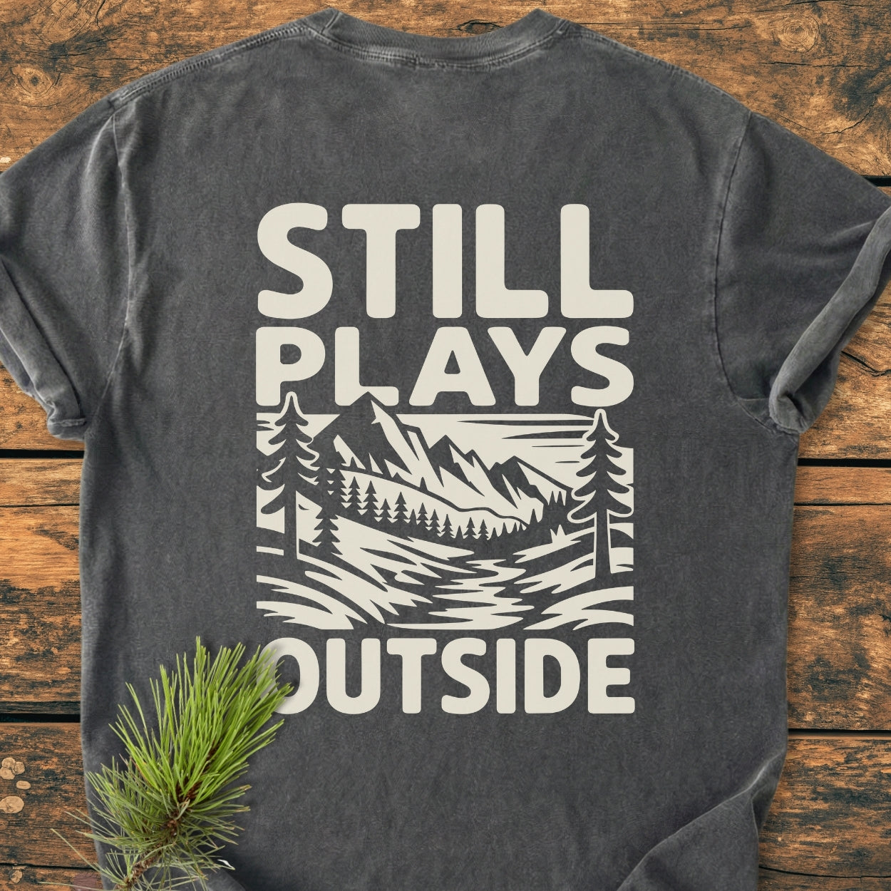 Charcoal-gray Still Plays Outside BACK PRINT - Vintage Wash Tee with a cream mountain and forest graphic on the back, laid on wood. Pine branch enhances the outdoor theme.
