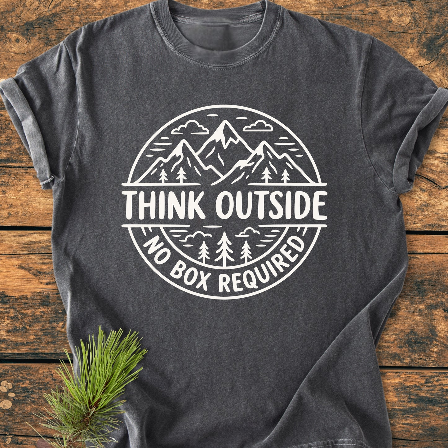 Think Outside - Vintage Wash Tee features a charcoal-gray design with a white outdoor-themed graphic, showcasing mountains and trees. Laid flat on wood, it includes a pine twig for a rustic touch.