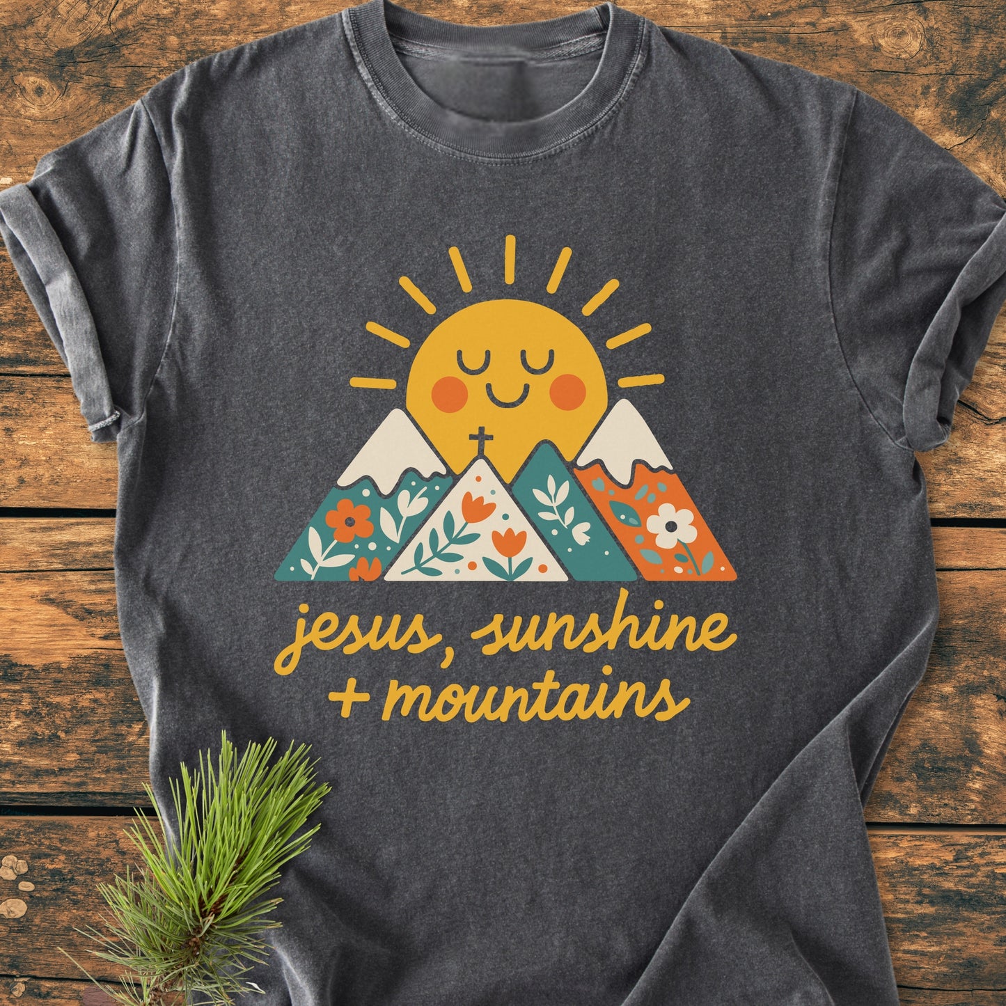 Jesus, Sunshine, Mountains - Vintage Heavyweight Tee featuring a smiling sun and floral-patterned mountains graphic, laid flat, showcasing its relaxed fit and durable double-needle stitching on soft charcoal-gray fabric.