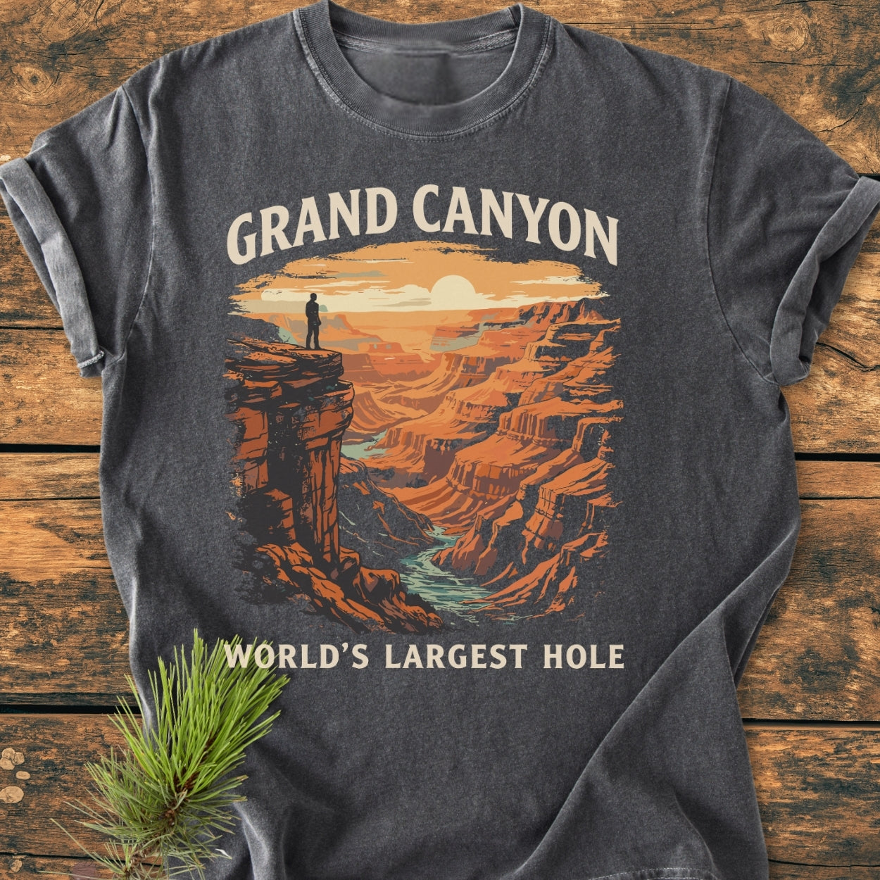 Grand Canyon Large Hole - Vintage Wash Tee features a sunset illustration of the Grand Canyon with a lone hiker, printed on a dark-gray, short-sleeve T-shirt laid on a rustic wooden surface.