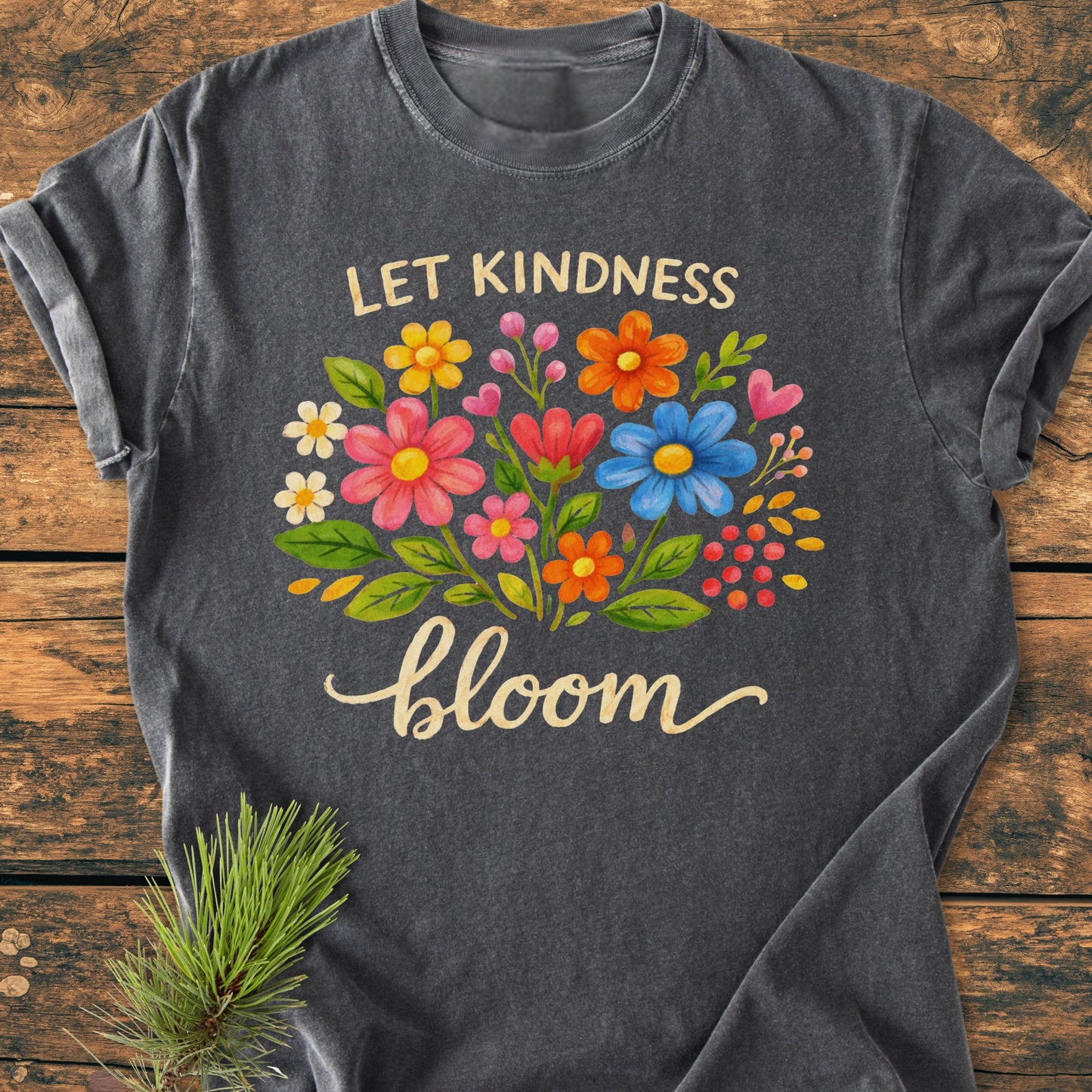 Let Kindness Bloom - Vintage Wash Tee featuring a vibrant floral graphic and whimsical text, laid flat on wood. Made from soft, durable, pre-shrunk cotton, offering a relaxed fit.