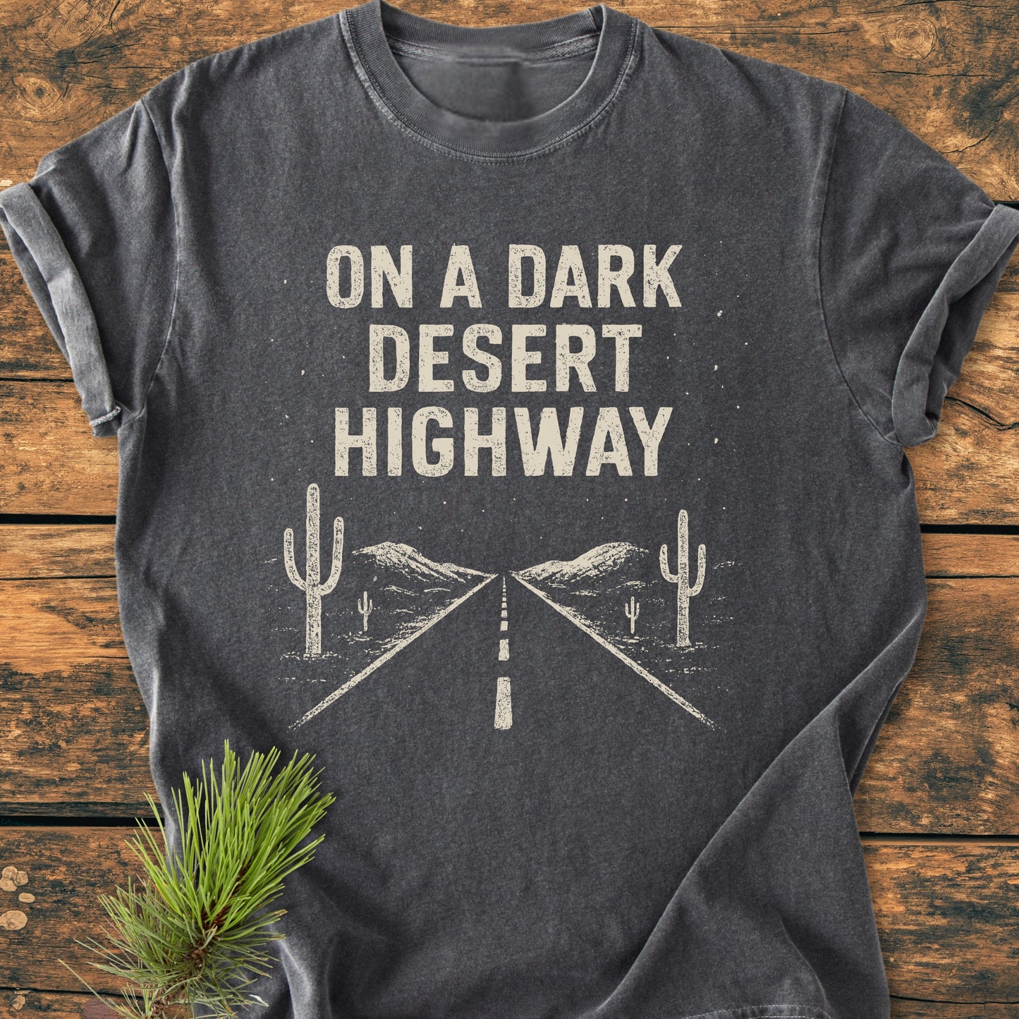 On A Dark Desert Highway - Vintage Wash Tee features a distressed graphic of a highway, cacti, and mountains on a charcoal-gray short-sleeve shirt, laid flat with pine sprigs.