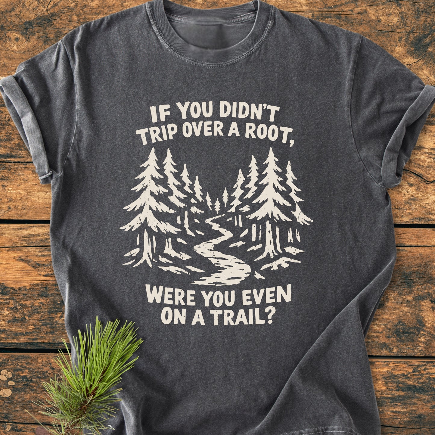 Were You Even On A Trail? - Vintage Wash Tee features a pine tree and trail graphic with a humorous slogan on a charcoal-gray T-shirt, accented by a pine sprig on a wooden backdrop.