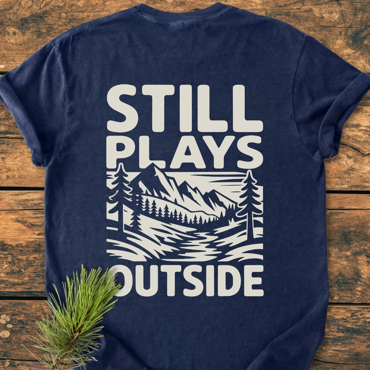 The Still Plays Outside BACK PRINT - Vintage Wash Tee showcases a white outdoor scene design on a dark-blue T-shirt, displayed on wood, with a pine sprig accentuating its natural theme.