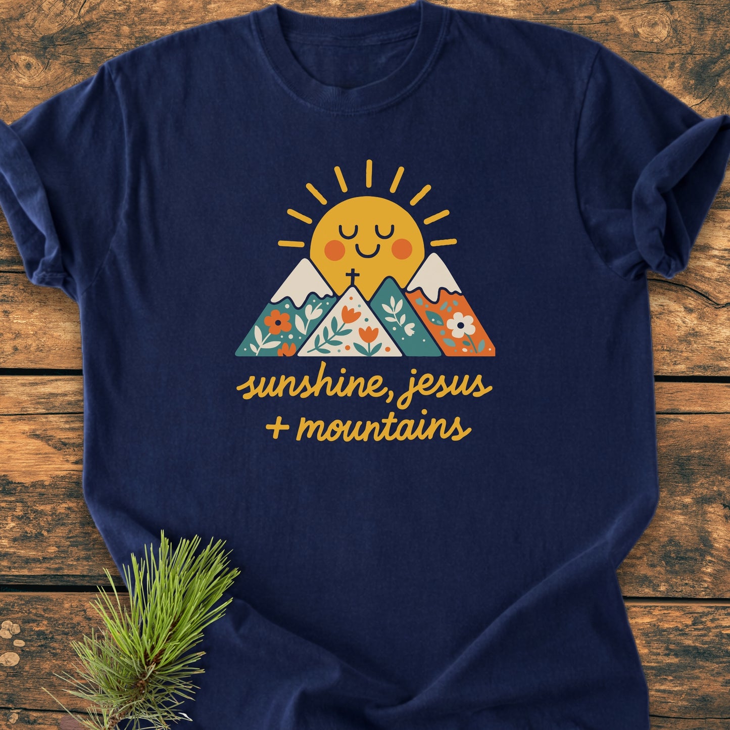 Jesus, Sunshine, Mountains - Vintage Heavyweight Tee features a smiling sun behind mountains with floral designs and a cross. Includes sunshine, jesus + mountains text. Laid on rustic wood with pine bough accent.