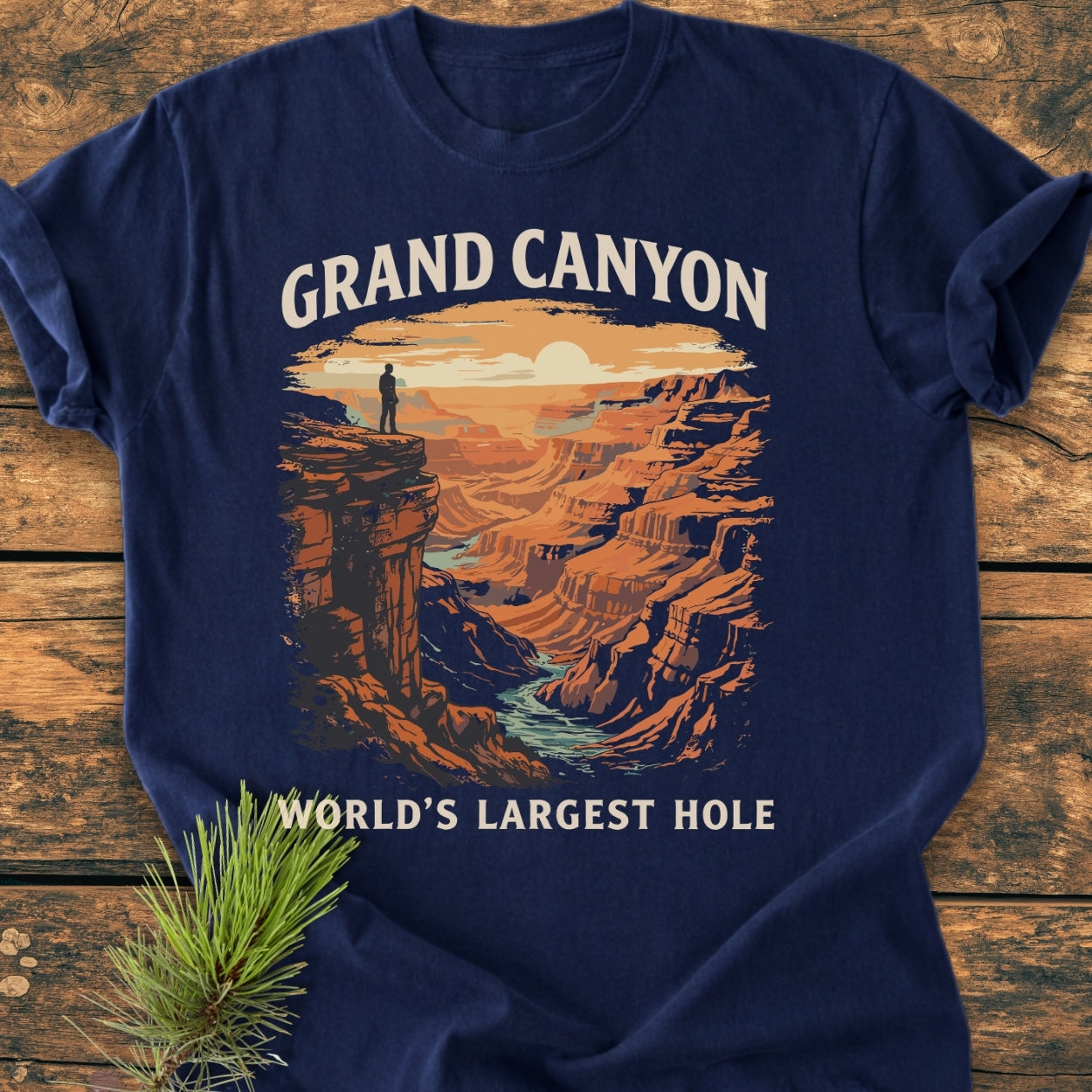 Grand Canyon Large Hole - Vintage Wash Tee features a stylized sunset Grand Canyon print, with a lone figure on a cliff, laid on a wooden surface, alongside a pine sprig.