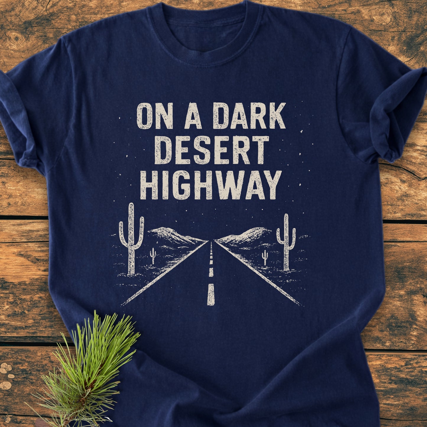 On A Dark Desert Highway - Vintage Wash Tee with distressed lettering and road graphic, laid flat on wood. Pine needles accent the shirt, embodying a trail lover's dream.