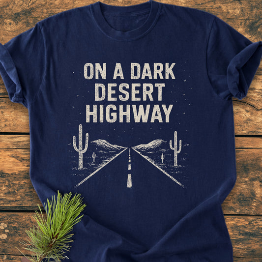 On A Dark Desert Highway - Vintage Wash Tee with distressed lettering and road graphic, laid flat on wood. Pine needles accent the shirt, embodying a trail lover's dream.