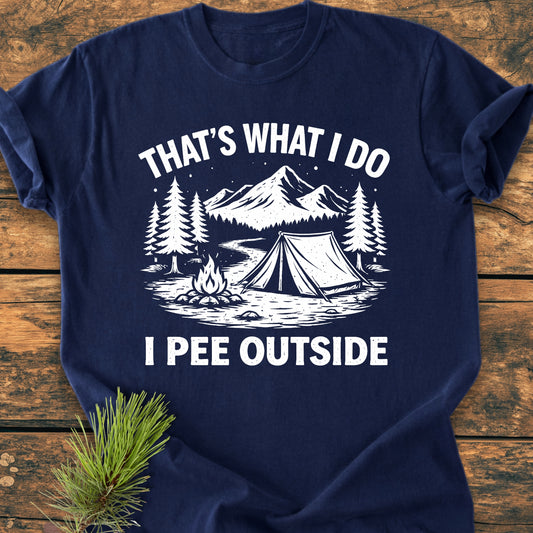 I Pee Outside - Vintage Wash Tee features a white camping scene with mountains, pine trees, tent, river, and campfire on a navy-blue background, laid on a wooden surface with a pine branch prop.