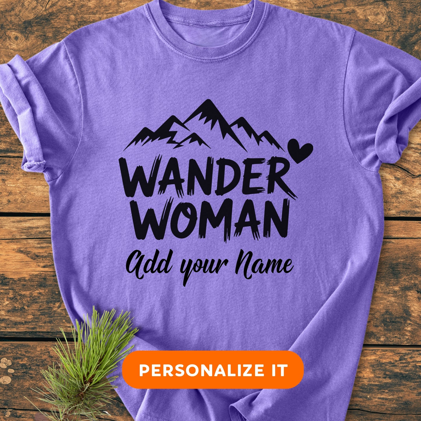 Lavender T-shirt with WANDER WOMAN graphic, mountain peaks, and customizable text option. Lays on wood surface with pine sprig, PERSONALIZE IT button displayed, embodying the personalized vintage wash tee theme.