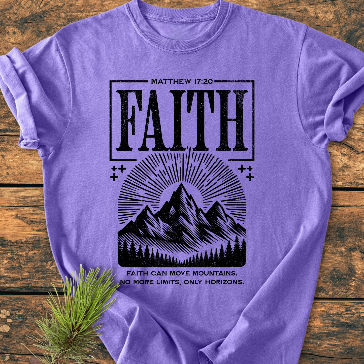 Faith Moves Mountains - Vintage Wash Tee features a black distressed graphic with 'FAITH' and mountain design on a purple T-shirt, laid flat on a wooden surface with a pine branch.