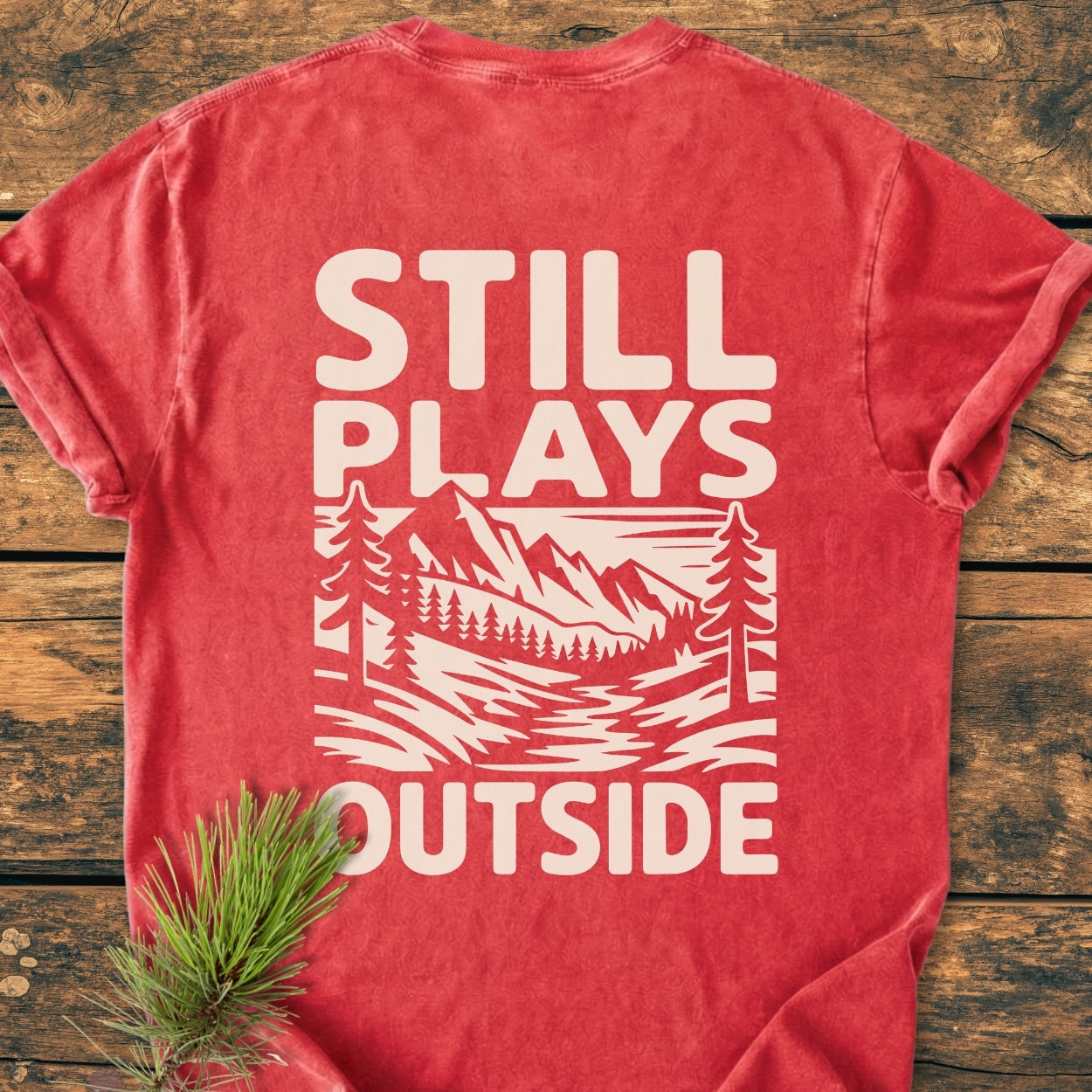 Still Plays Outside BACK PRINT - Vintage Wash Tee features a stylized outdoor scene on a red t-shirt, laid flat, highlighting pine trees and mountains, emphasizing comfort and durability in its design.