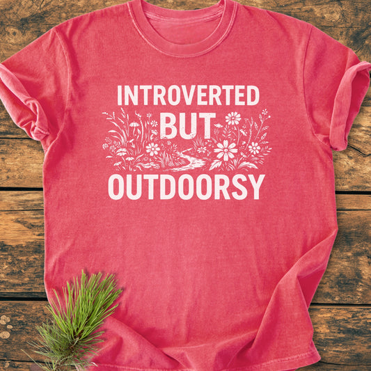 Introverted But Outdoorsy - Vintage Wash Tee features bold white text and a decorative wildflower graphic on a heathered reddish-pink T-shirt, laid flat on a wooden surface.