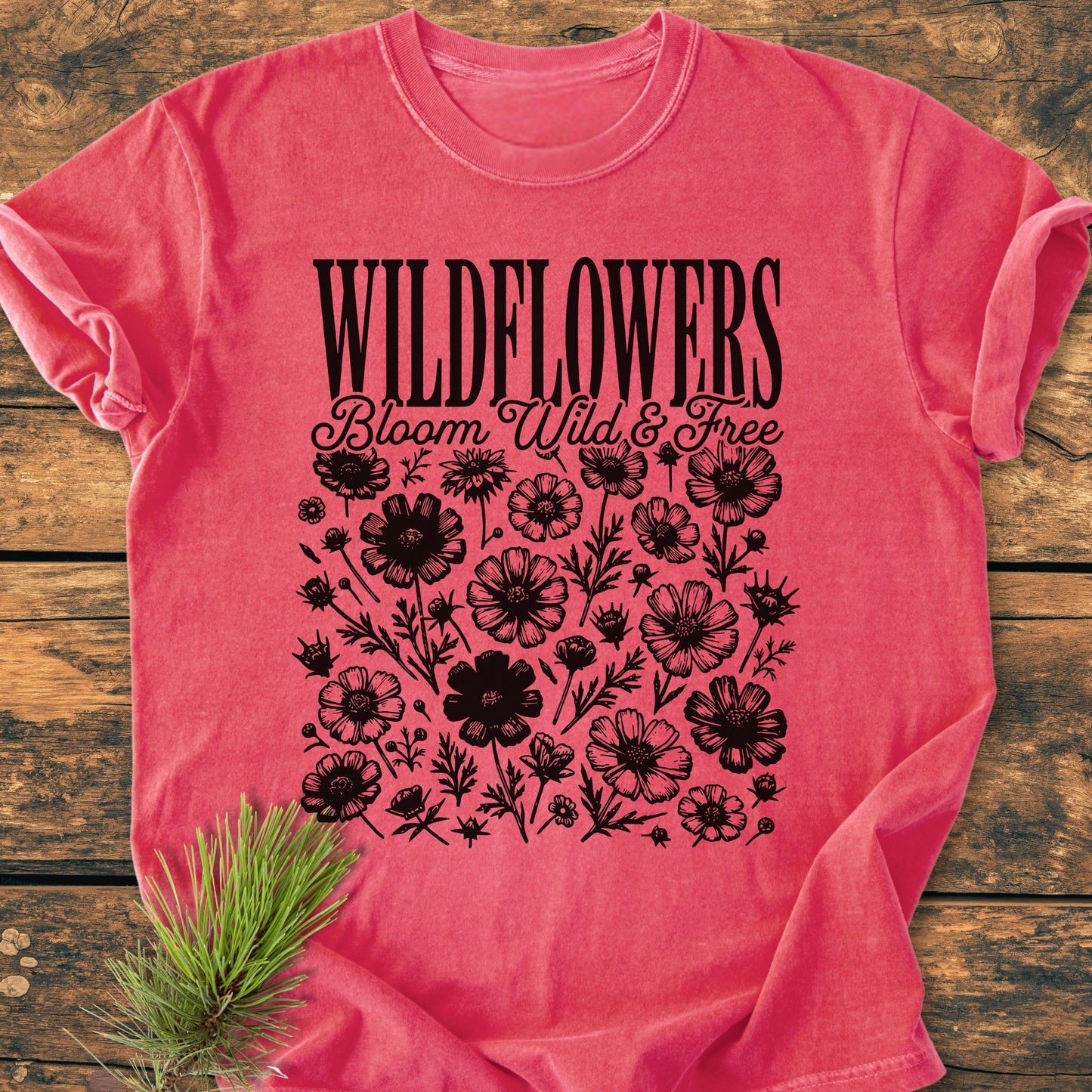 Wildflowers Bloom Wild And Free - Vintage Wash Tee displayed flat with black wildflower prints and styled text. A small fir-pine sprig accents the rustic presentation of this relaxed-fit, pre-shrunk cotton T-shirt.