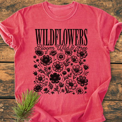 Wildflowers Bloom Wild And Free - Vintage Wash Tee displayed flat with black wildflower prints and styled text. A small fir-pine sprig accents the rustic presentation of this relaxed-fit, pre-shrunk cotton T-shirt.