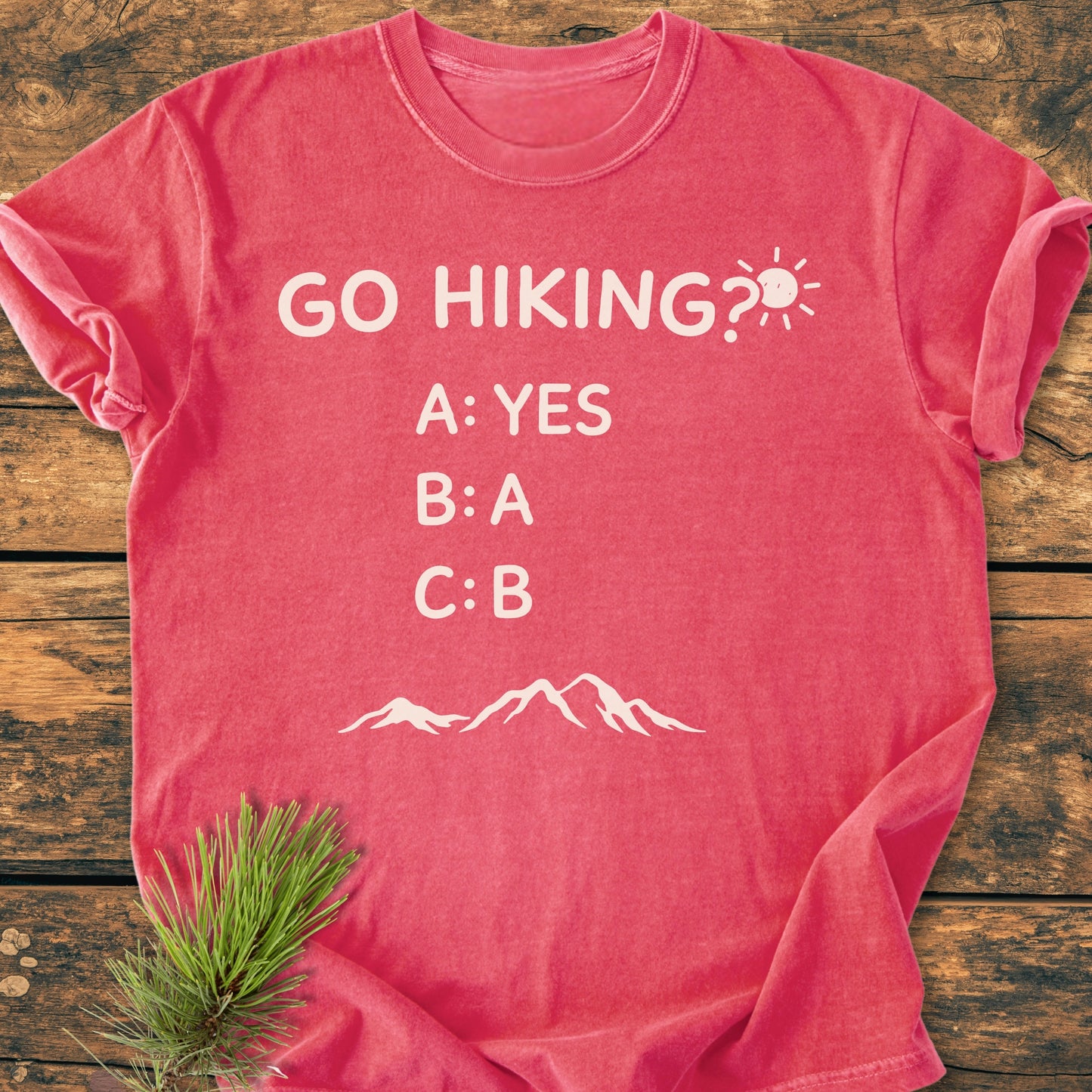 Go Hiking? - Vintage Wash Tee in heather-red, laid flat on wood. Features playful slogan with mountain silhouette, highlighting its relaxed fit and cozy, garment-dyed cotton fabric.