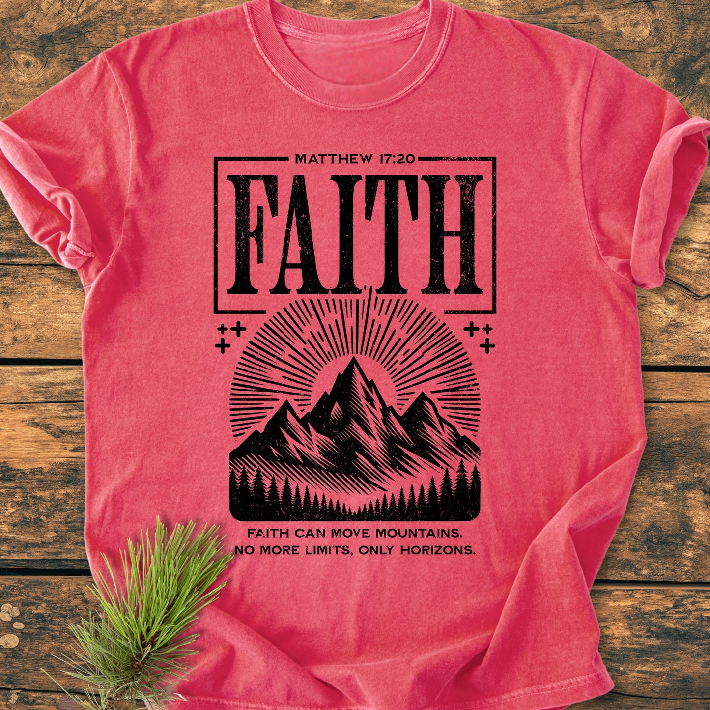 Faith Moves Mountains - Vintage Wash Tee, featuring a distressed FAITH design with mountain graphics and inspirational text, laid flat on a wooden surface, styled with a pine branch beside it.