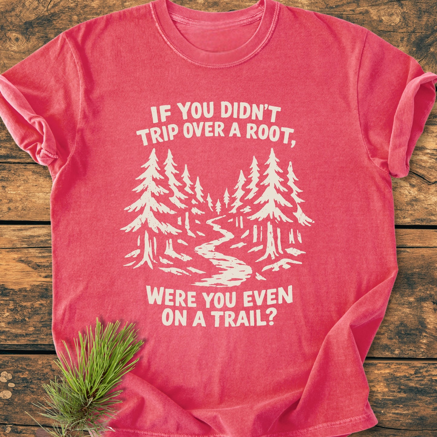 Were You Even On A Trail? - Vintage Wash Tee features a reddish-pink color with a forest scene and slogan, laid on wood, accented by pine needles.