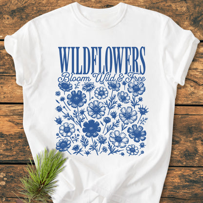 Wildflowers Bloom Wild And Free - Vintage Wash Tee features WILDFLOWERS in blue serif font, with a script tagline and wildflower illustrations, laid on wood with a pine sprig accent.