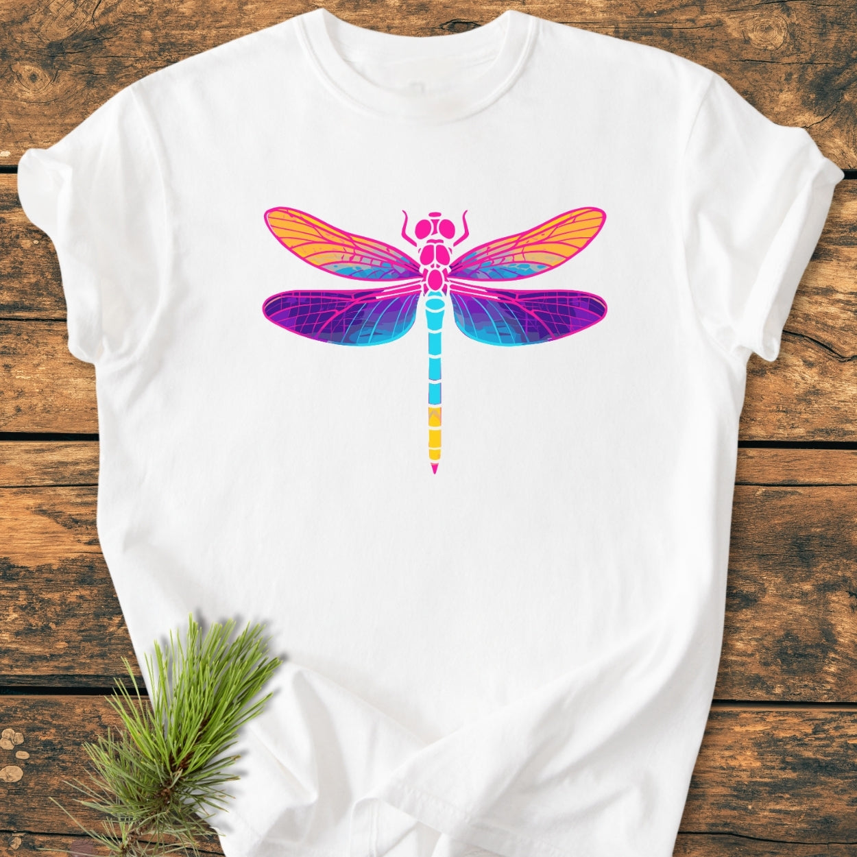 The Psychedelic Dragonfly - Vintage Wash Tee features a vibrant dragonfly graphic on a white, relaxed-fit, garment-dyed T-shirt, set against a rustic wooden background with pine needles accentuating the scene.