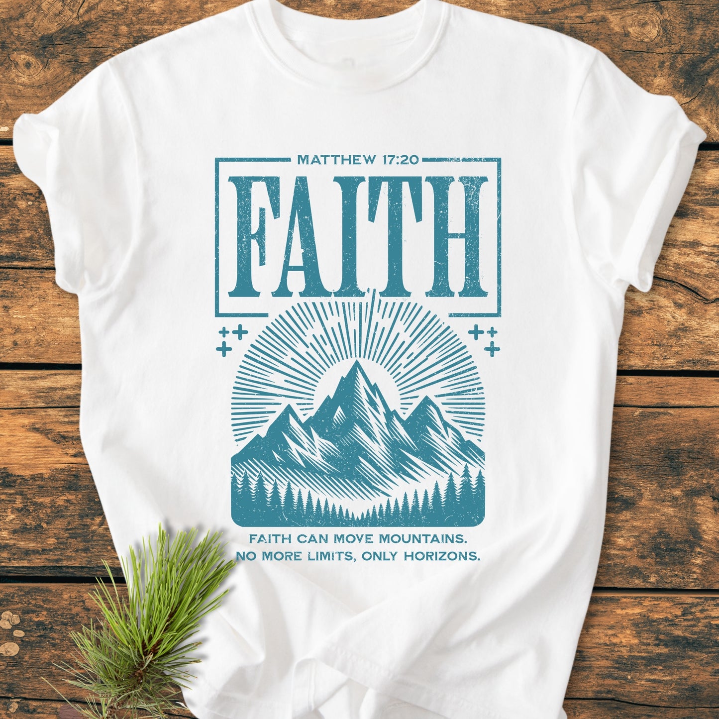 Faith Moves Mountains - Vintage Wash Tee: White cotton T-shirt with MATTHEW 17:20 and Faith can move mountains slogan, distressed teal graphic of sun-rays over mountains, laid on wooden surface with pine sprig.