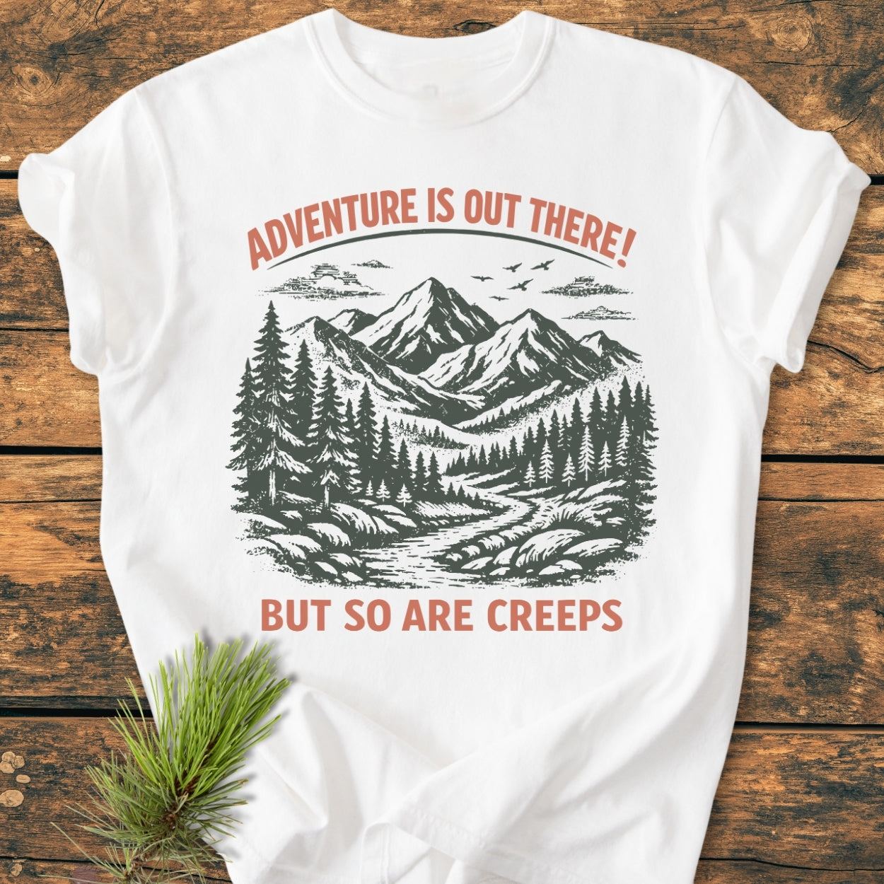 Adventure Is Out There - Vintage Wash Tee featuring a stylized mountain-and-forest scene with text overlay, laid on a wooden surface, complemented by a pine sprig, emphasizing its outdoor theme.