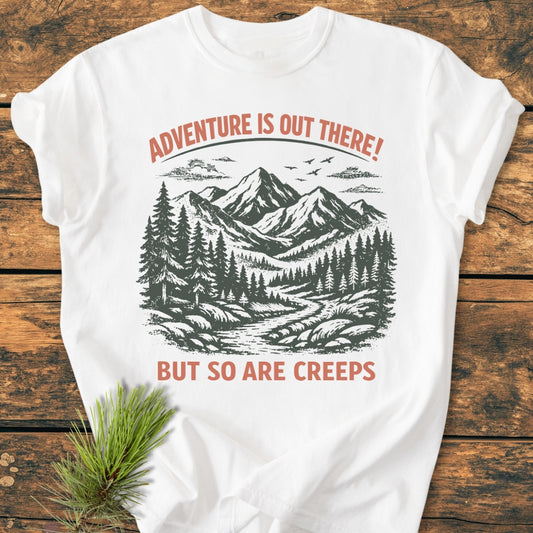 Adventure Is Out There - Vintage Wash Tee featuring a stylized mountain-and-forest scene with text overlay, laid on a wooden surface, complemented by a pine sprig, emphasizing its outdoor theme.